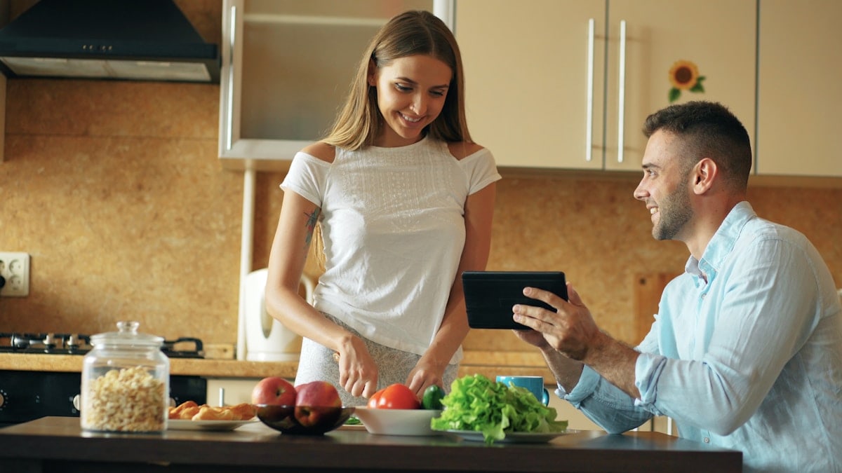 Attractive couple chatting in the kitchen early morning. Handsome man using tablet while his girlfriend cooking breakfast at home